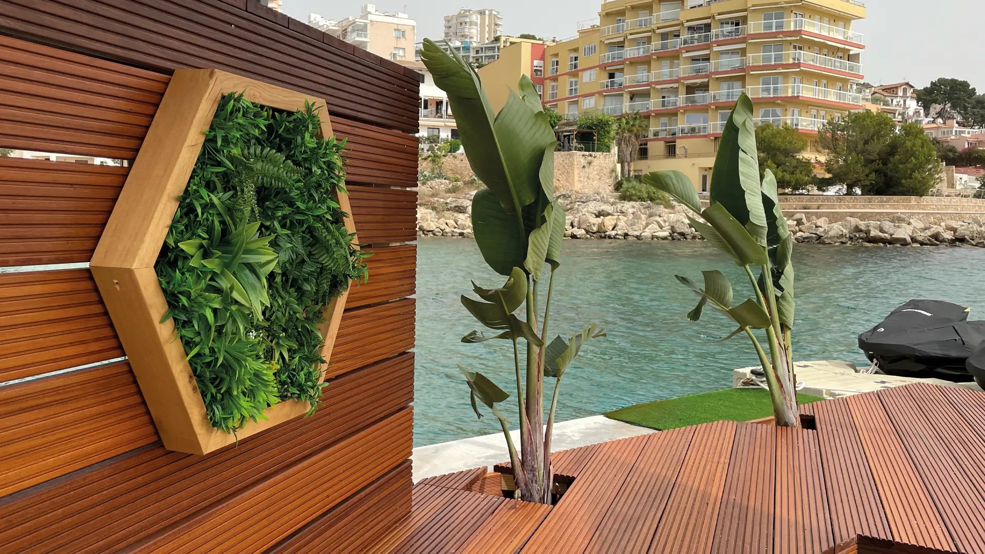 Sechseckiger grüner Wandpflanzer auf Holzdeck mit Blick aufs Wasser.