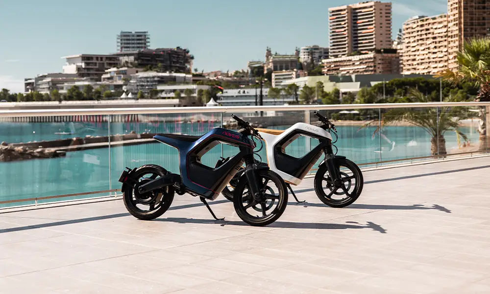 Zwei Novus-Bikes stehen nebeneinander, das linke ist blau und das rechte ist weiß und golden, während das Meer und eine Stadt im Hintergrund zu sehen sind.