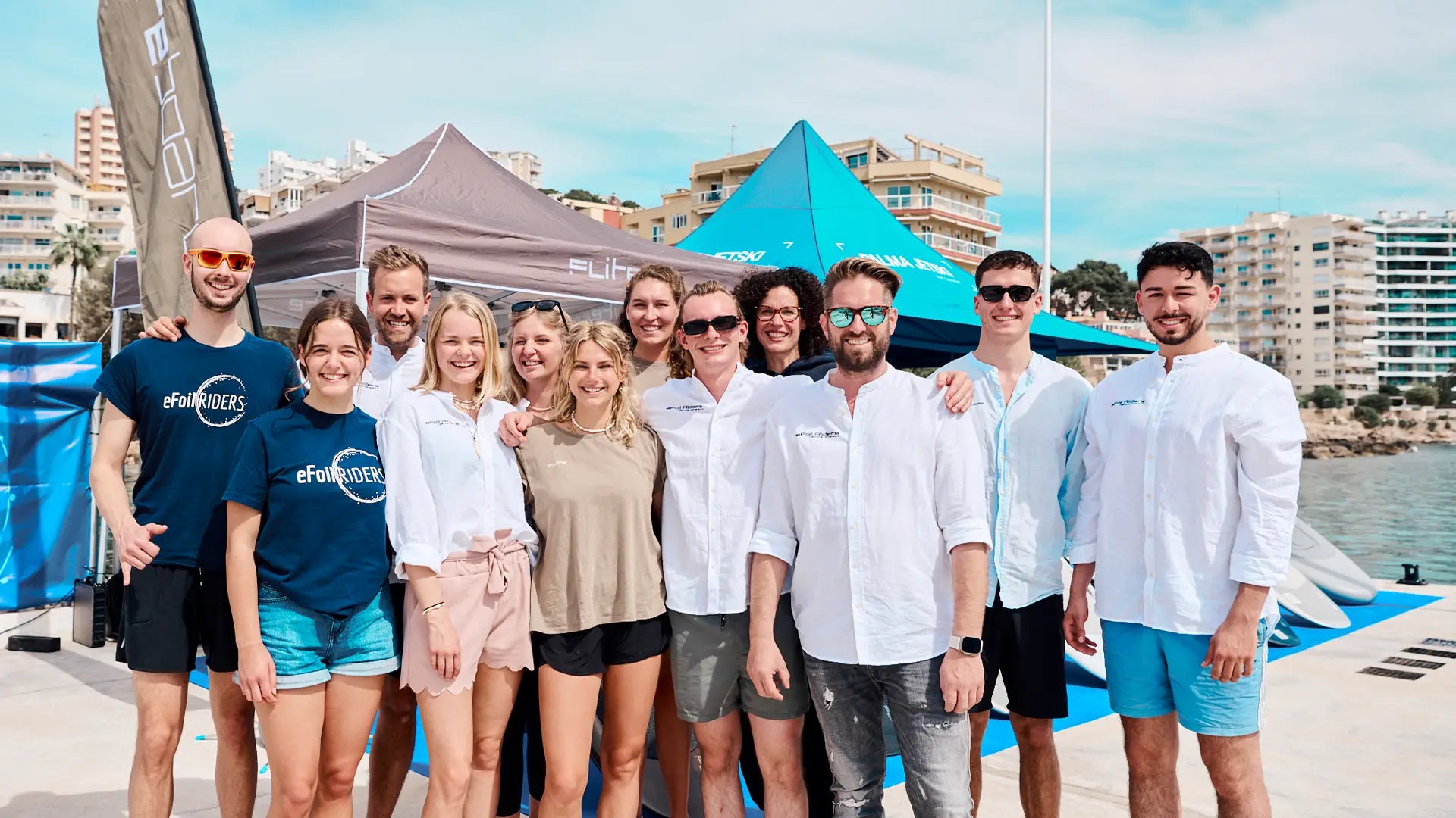 Eine Gruppe von Leuten posiert für ein Foto an einem Ufer mit Zelten und Gebäuden im Hintergrund.