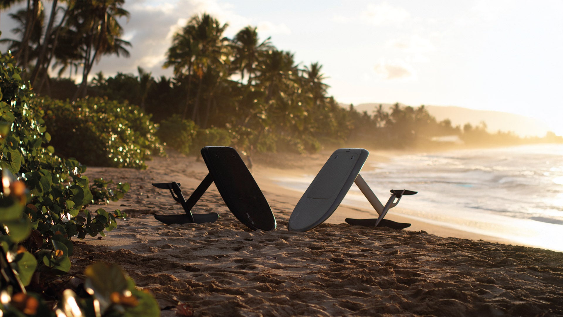 Zwei Fliteboards, schwarz auf der linken und weiß auf der rechten Seite, sind am Strand aufgestellt, während die Sonne im Hintergrund untergeht und die tropische Küste in ein schönes Licht taucht.