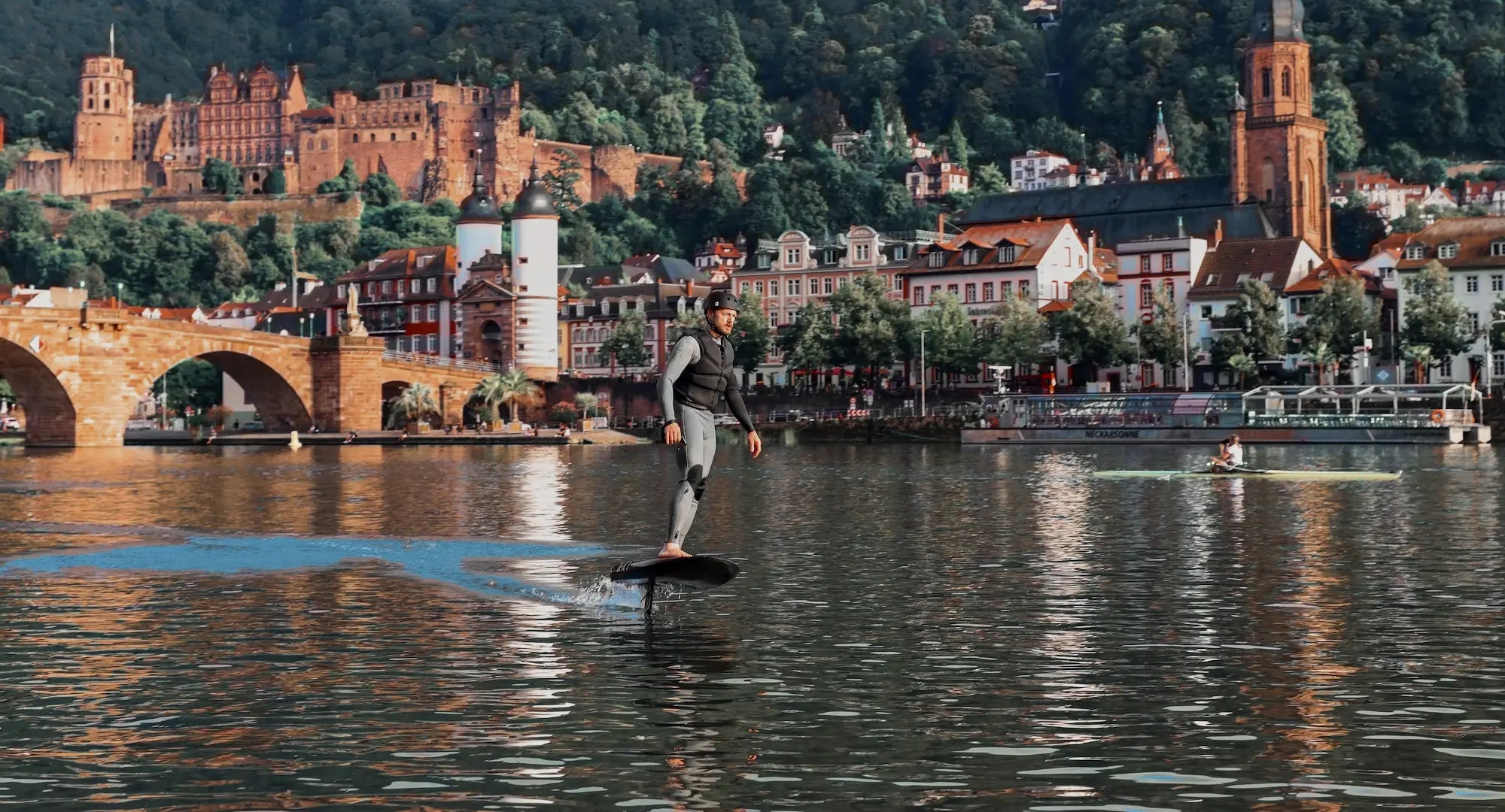 Ein Mann in Neoprenanzug und Helm fliegt auf seinem Fliteboard über den Fluss in Heidelberg, mit dem Schloss und der Stadt im Hintergrund.