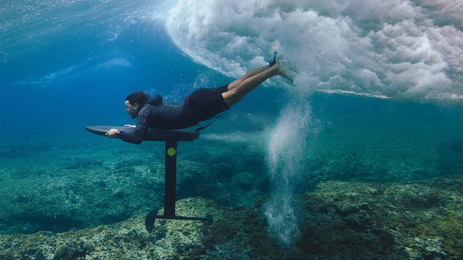 Person im Neoprenanzug, die auf einem FliteLab-Surfbrett unter Wasser liegt, mit einer großen Welle im Hintergrund.