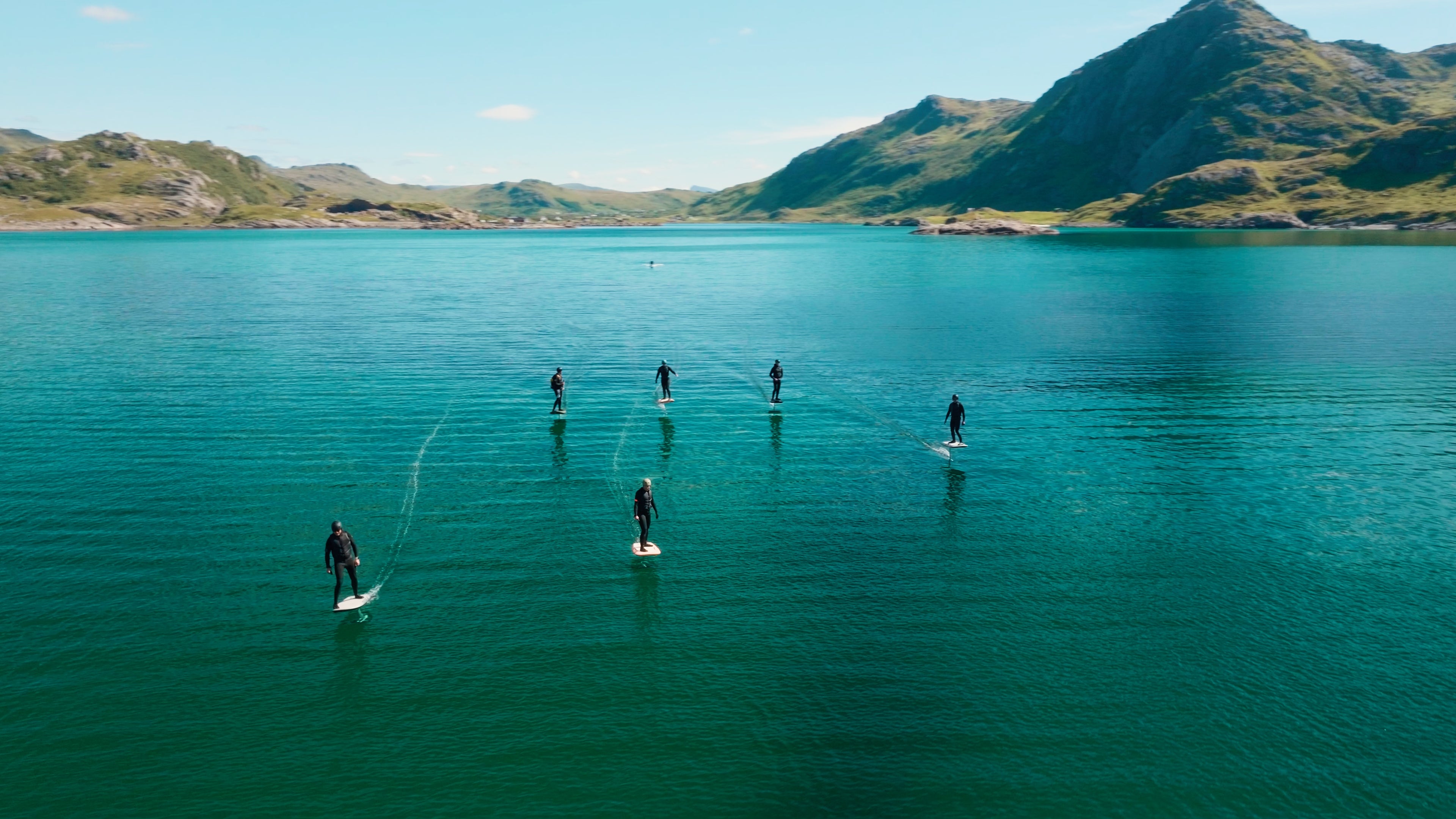 Sechs Personen eFoiling auf Fliteboards in Norwegen auf dem türkisfarbenen Wasser, im Hintergrund Du kannst eine schöne Landschaft mit Bergen sehen.
