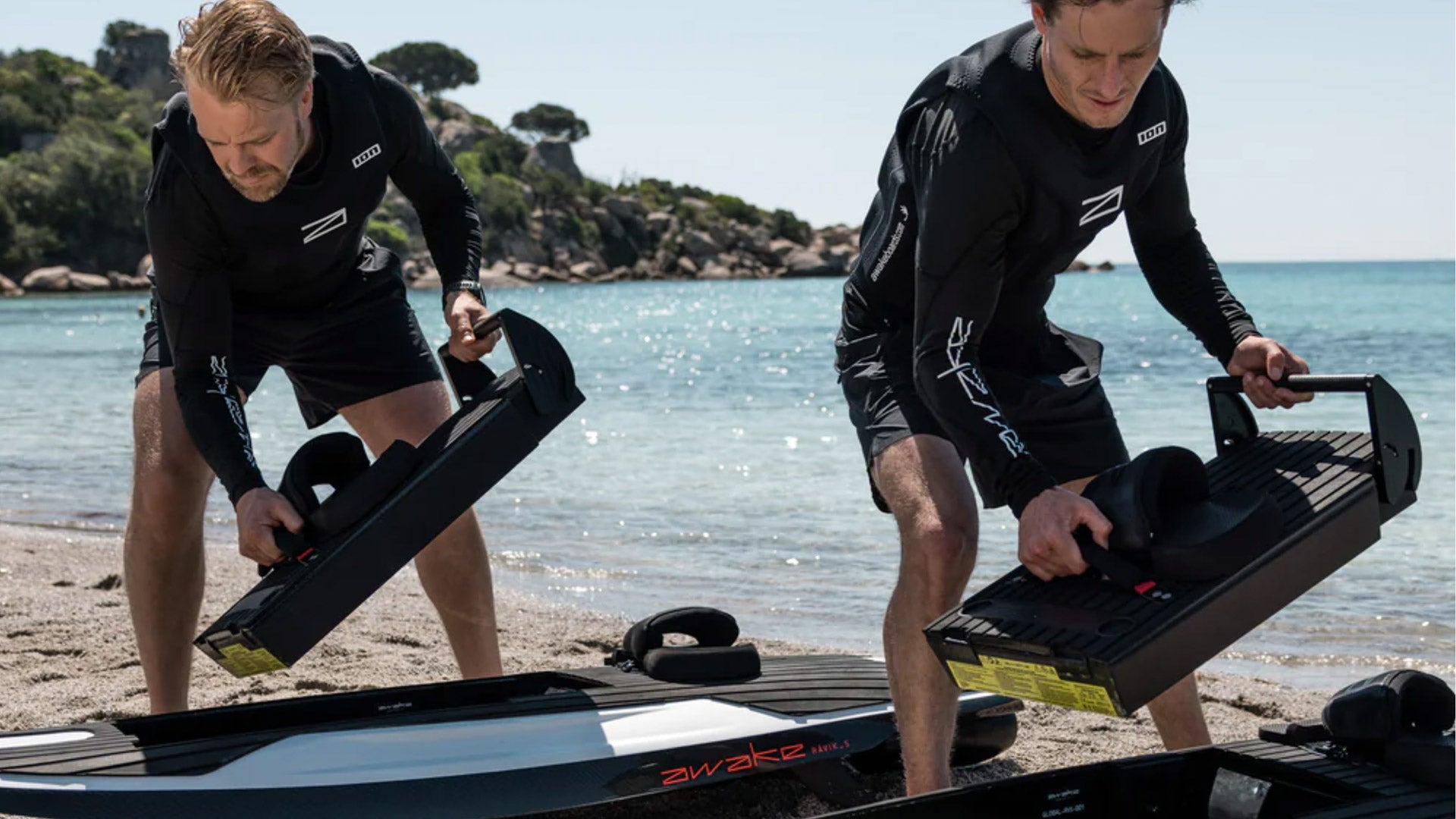 Zwei männliche Awake-Instruktoren stehen an einem Strand und setzen Batterien in die Awake-Boards ein.