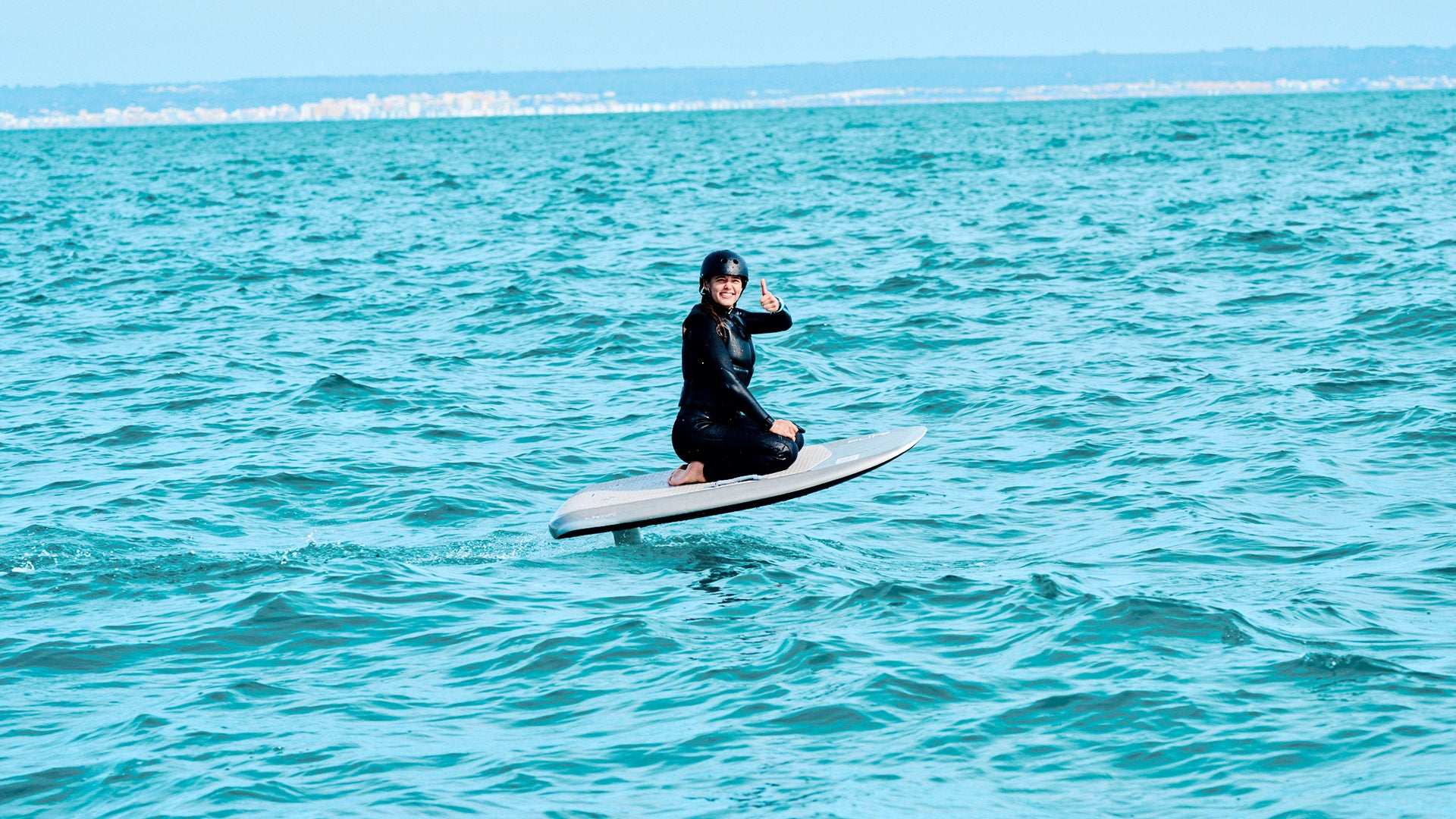 Eine Frau sitzt auf einem Fliteboard und schwebt über dem Meer, während sie mit dem Daumen nach oben in die Kamera zeigt.
