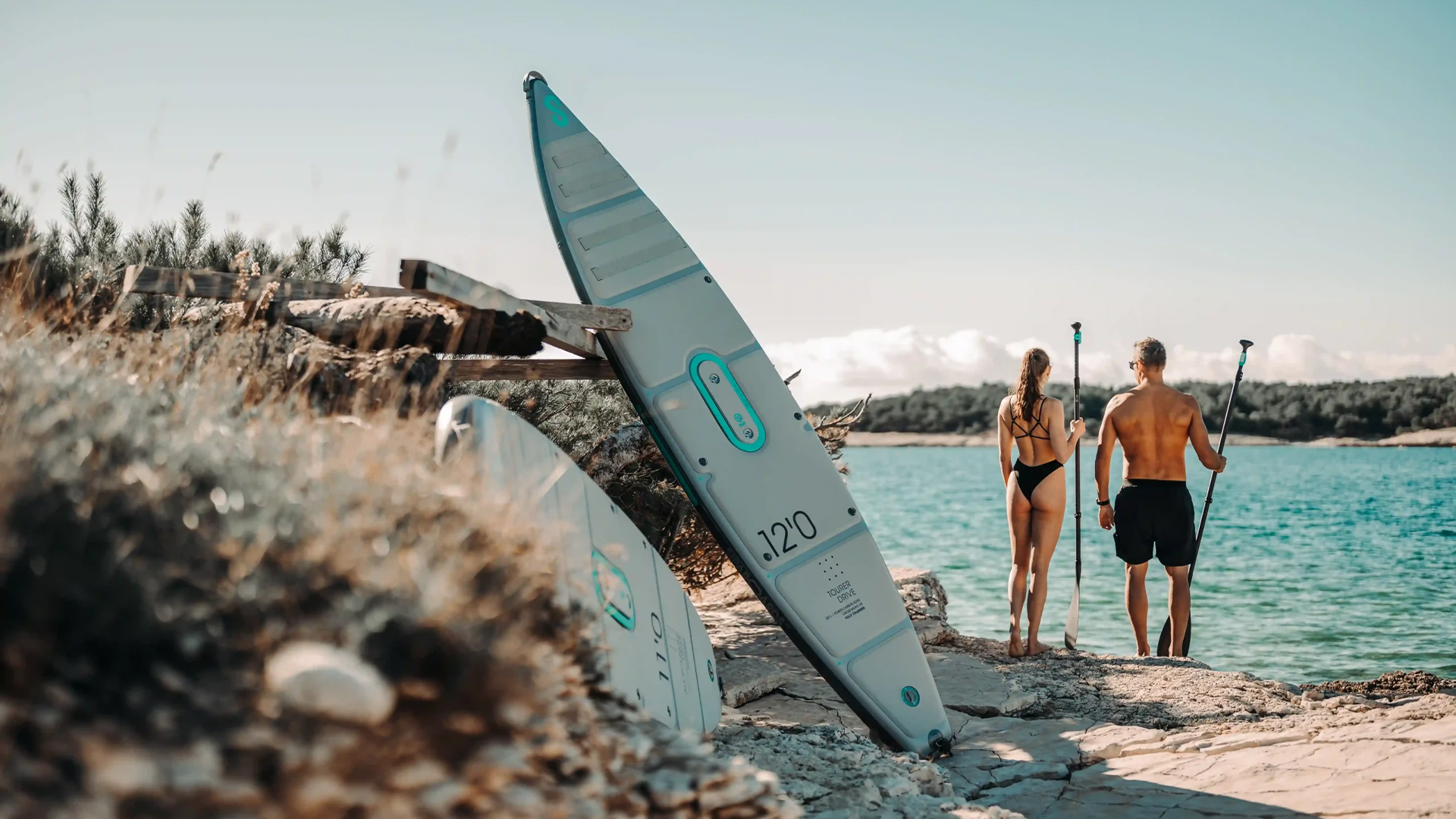Zwei Sipa-Boards lehnen am Holzbalken am Strand, während im Hintergrund eine Frau und ein Mann das Paddel halten und aufs Meer hinausschauen.