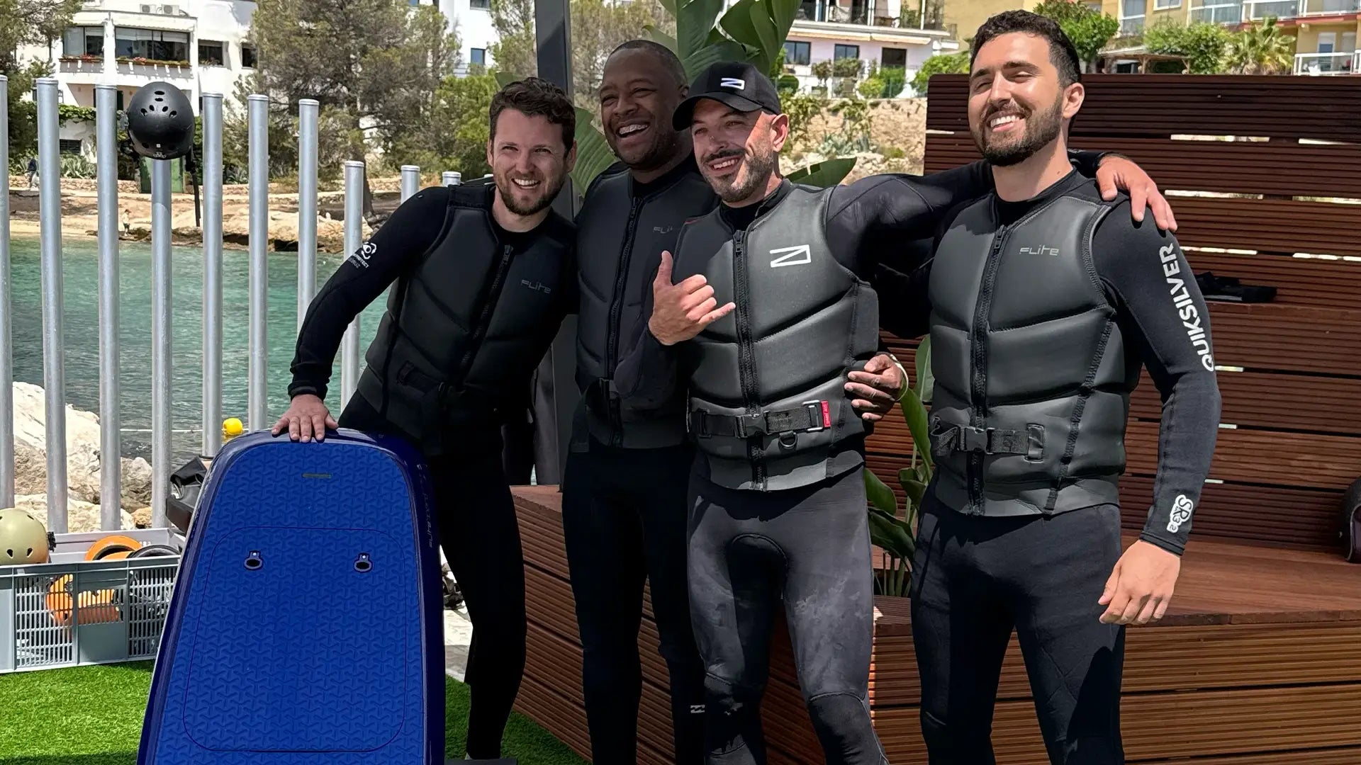 Vier Männer in Neoprenanzügen stehen zusammen an einem Ufer mit einem blauen Fliteboard.
