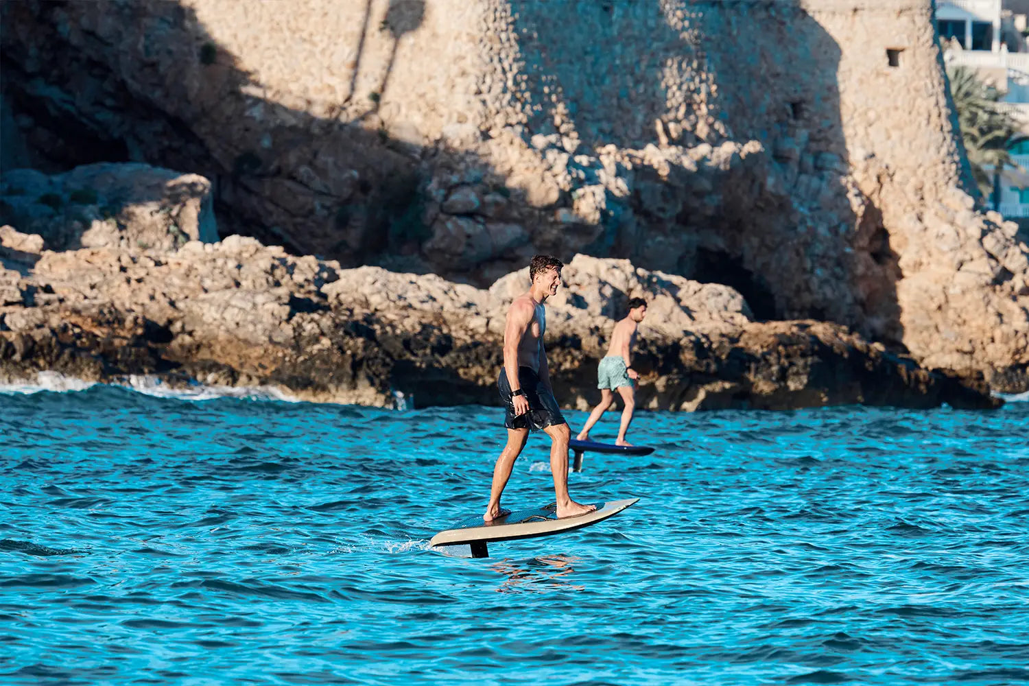 Zwei Männer fliegen auf einem Fliteboard über das Wasser in Mallorca, mit braunen Felsen im Hintergrund.