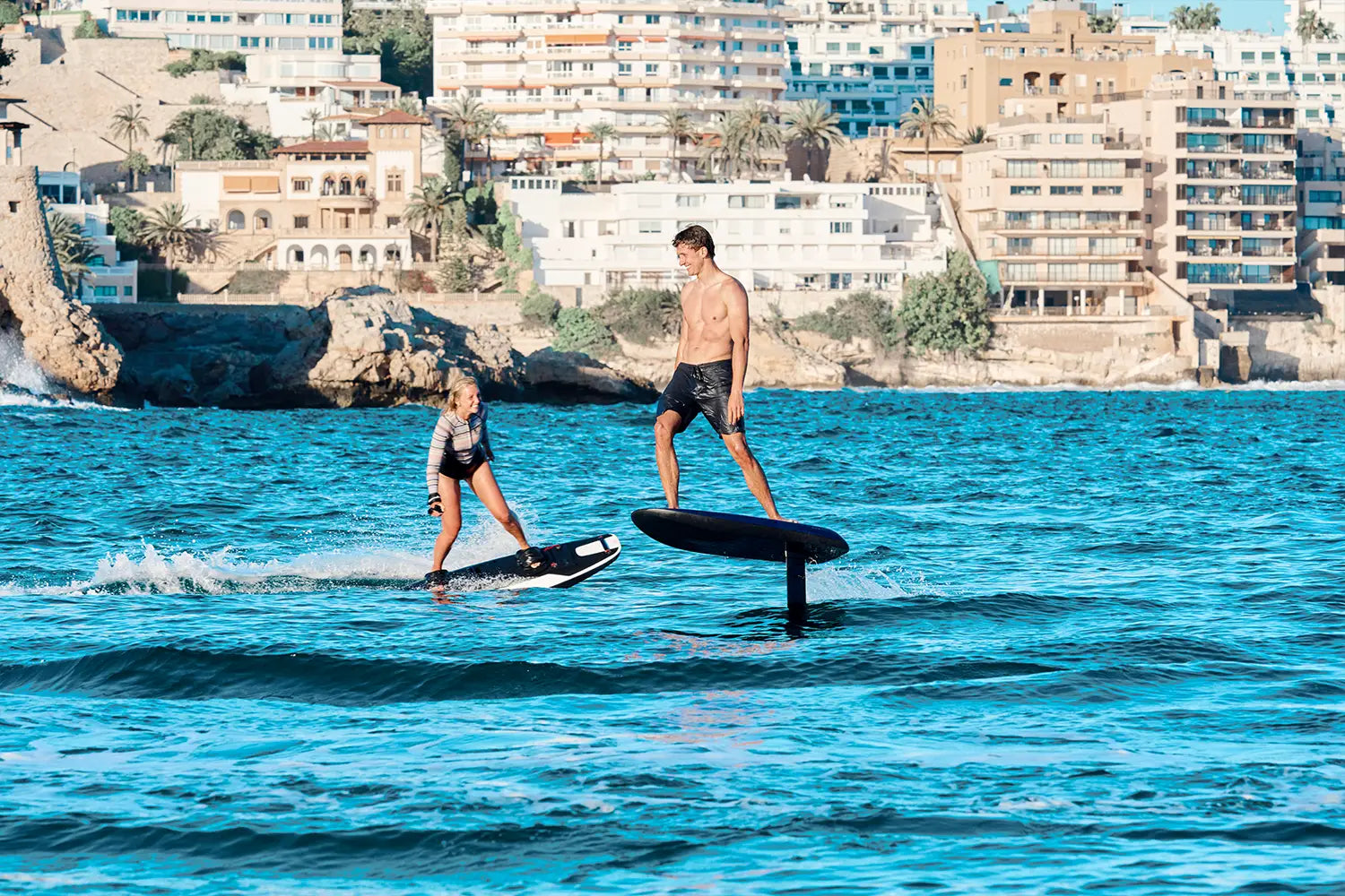 Ein Junge fährt glücklich auf einem Fliteboard auf dem Meer in Richtung einer Frau, die ein Awake Jetboard fährt, mit Mallorcas Küstenlinie im Hintergrund.
