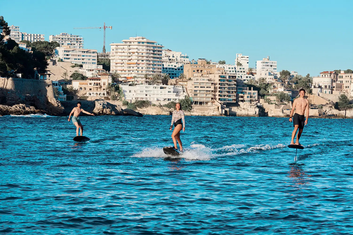 Rechts und links fahren zwei Männer mit einem Fliteboard auf dem Meer, in der Mitte fährt eine Frau mit einem Awake Jetboard, mit Mallorcas Küstenlinie im Hintergrund.