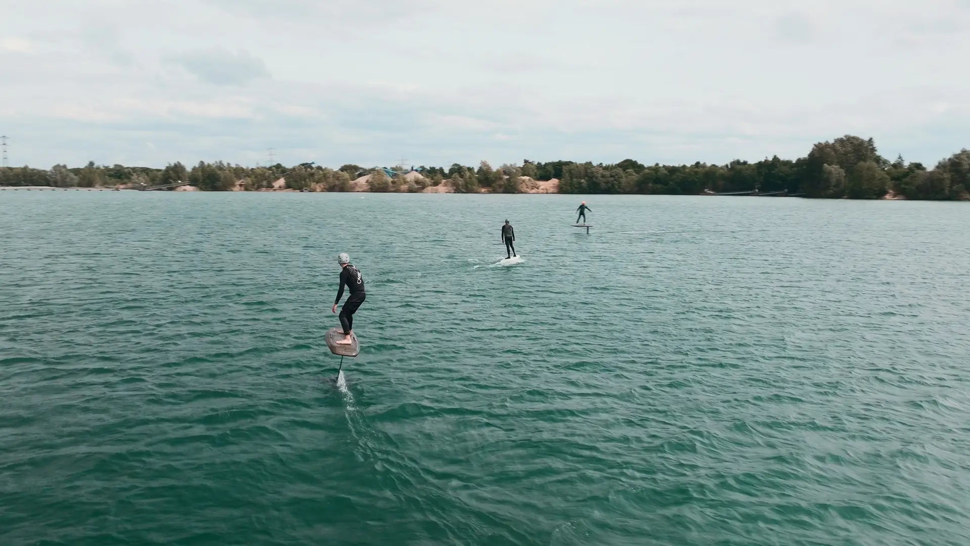 Drei Personen beim eFoilen auf einem ruhigen See mit Bäumen und Gebäuden im Hintergrund.