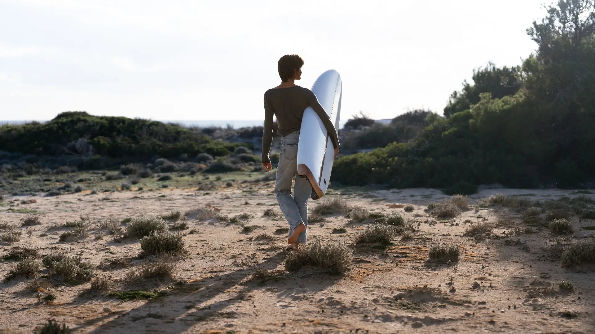 Eine Frau geht in Jeans und T-Shirt am Strand von der Kamera weg und hält ein Lind Surfboard unter dem Arm.