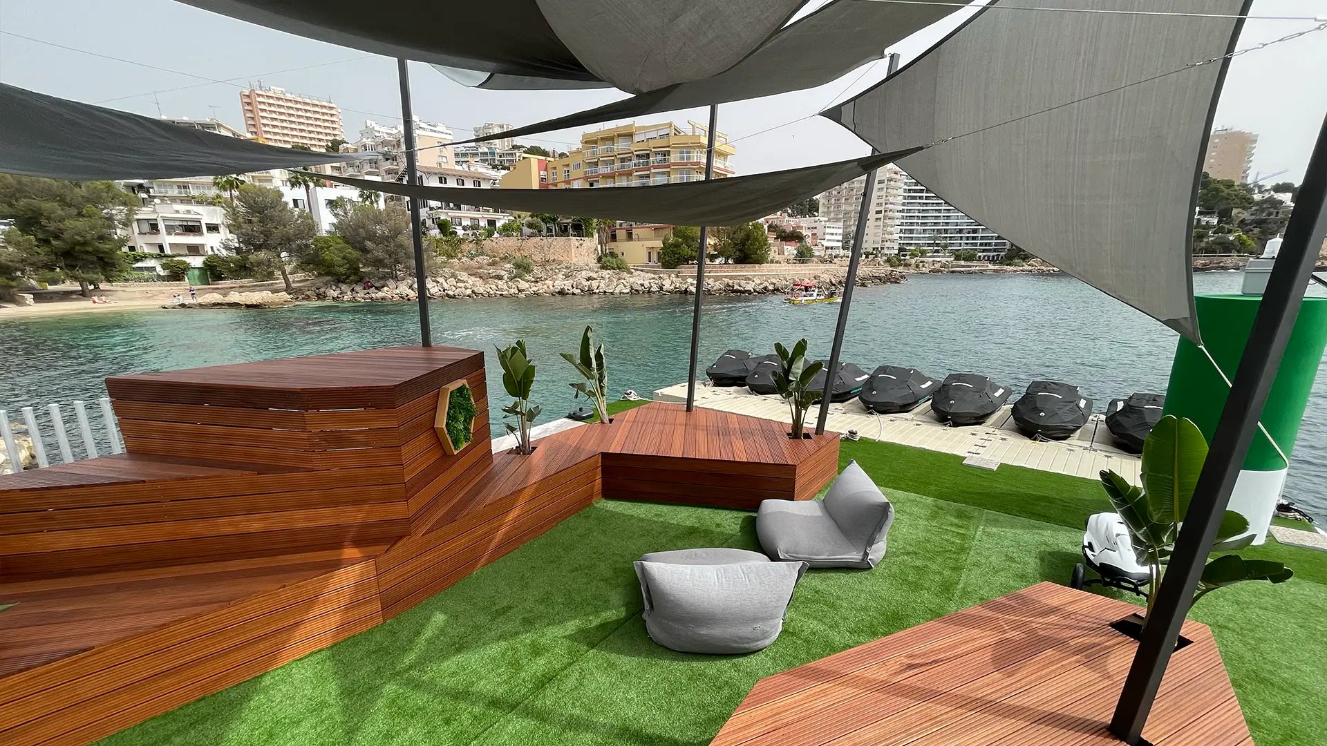 Sitzbereich im Freien mit Holzbänken und grauen Kissen an einem Ufer mit Stadtkulisse.