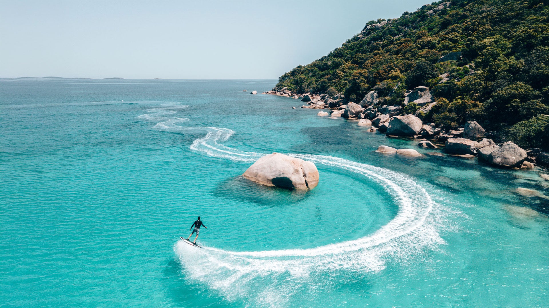 Eine Vogelperspektive einer Person, die mit einem Awake Raevik S Jetboard um einen Felsen auf dem Meer fährt.