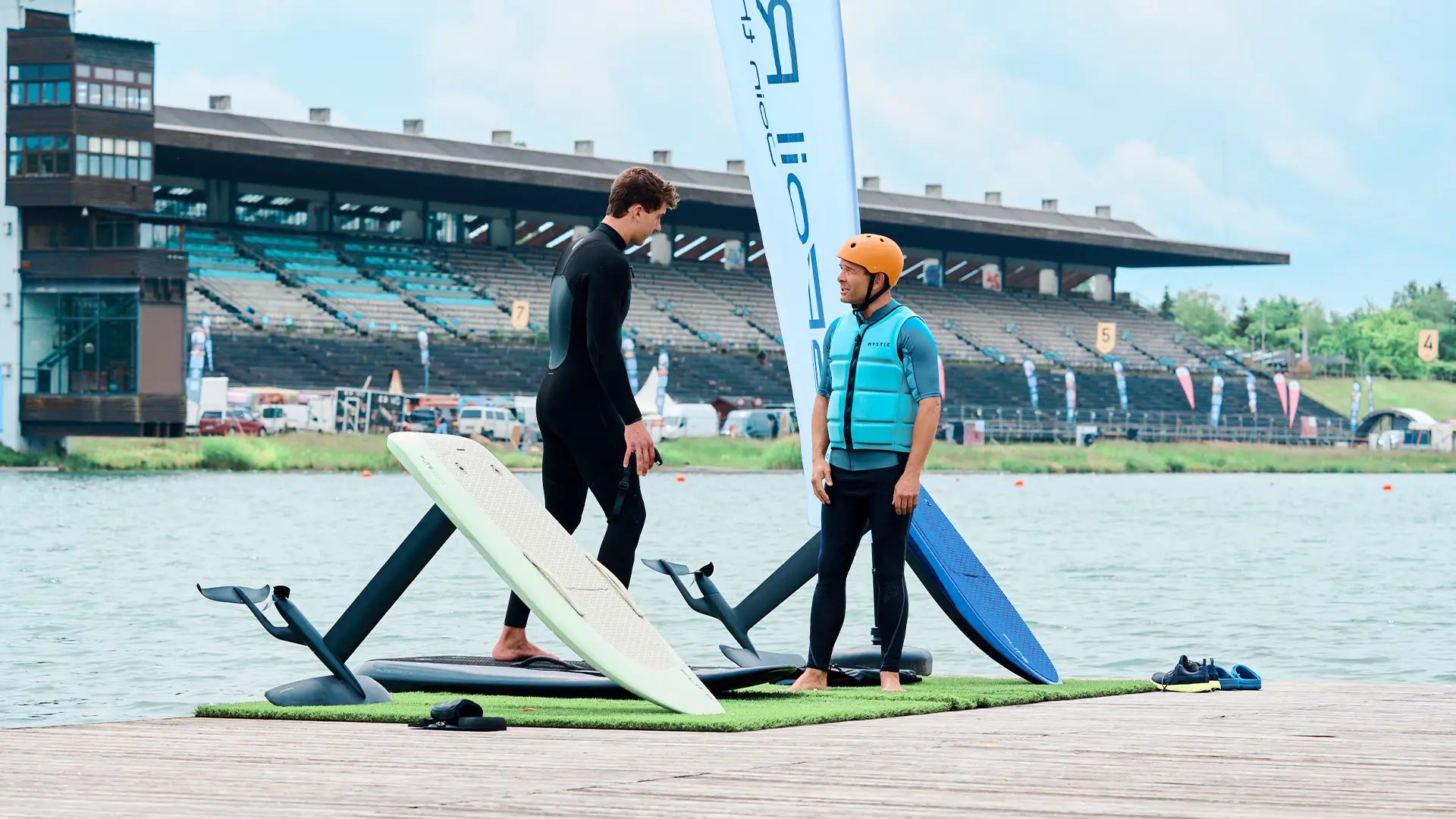 Ein Surflehrer erklärt seinem Schüler auf einem Pier, wie man ein Fliteboard fährt. Neben ihnen stehen verschiedene Boards und im Hintergrund sieht man die Regattastrecke in München.