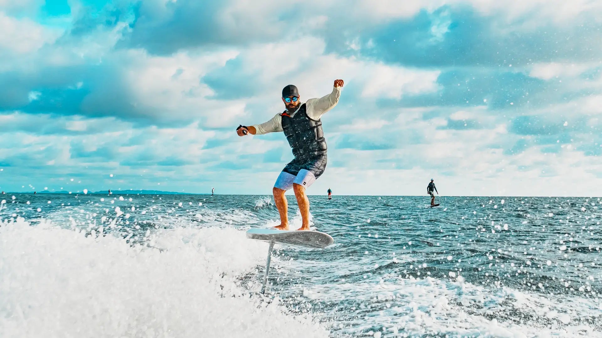 Ein Mann mit Sonnenbrille ist auf einem fliteboard auf dem Meer unterwegs, im Hintergrund sieht man andere Leute beim Segeln.