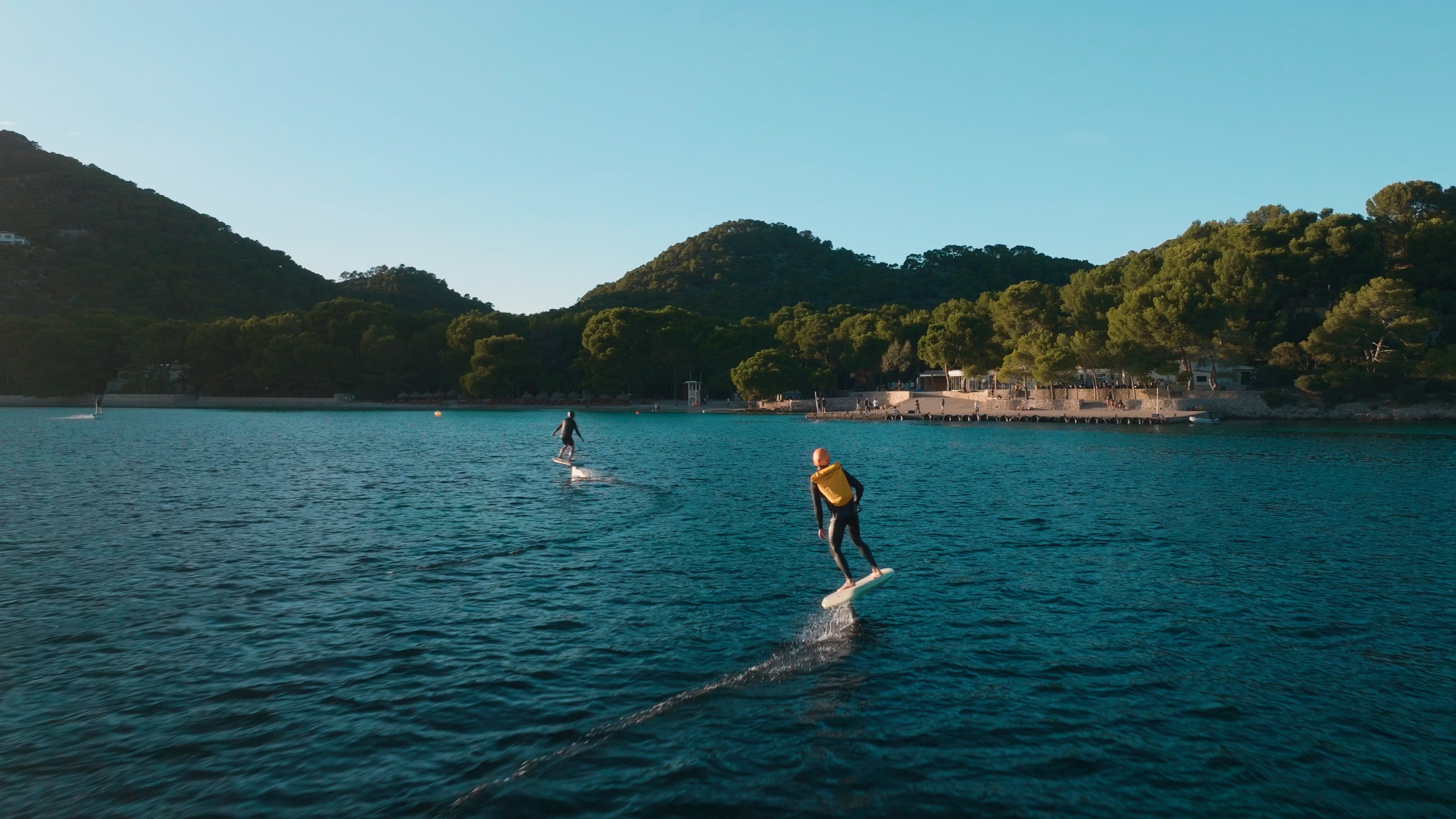 Zwei Personen treiben mit Surfbrettern auf dem Meer, im Hintergrund sind Berge mit grüner Vegetation Du kannst sehen.