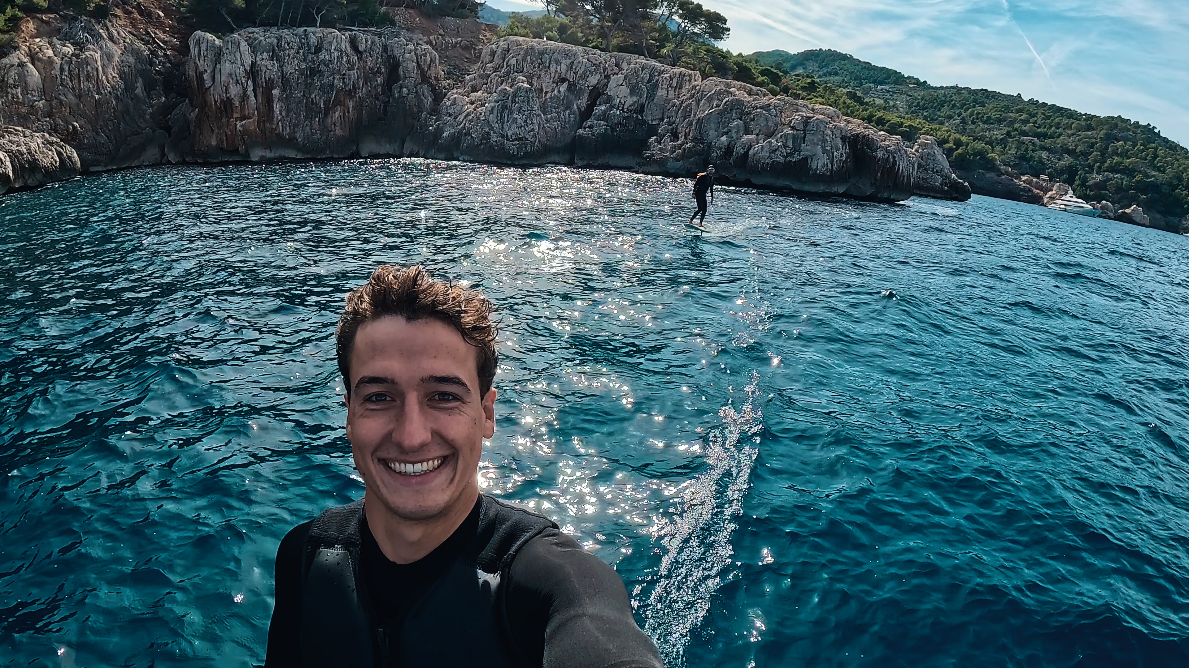 Eine Person auf dem Meer lächelt in die Kamera, während hinter ihm eine andere Person auf einem Fliteboard efoilt.