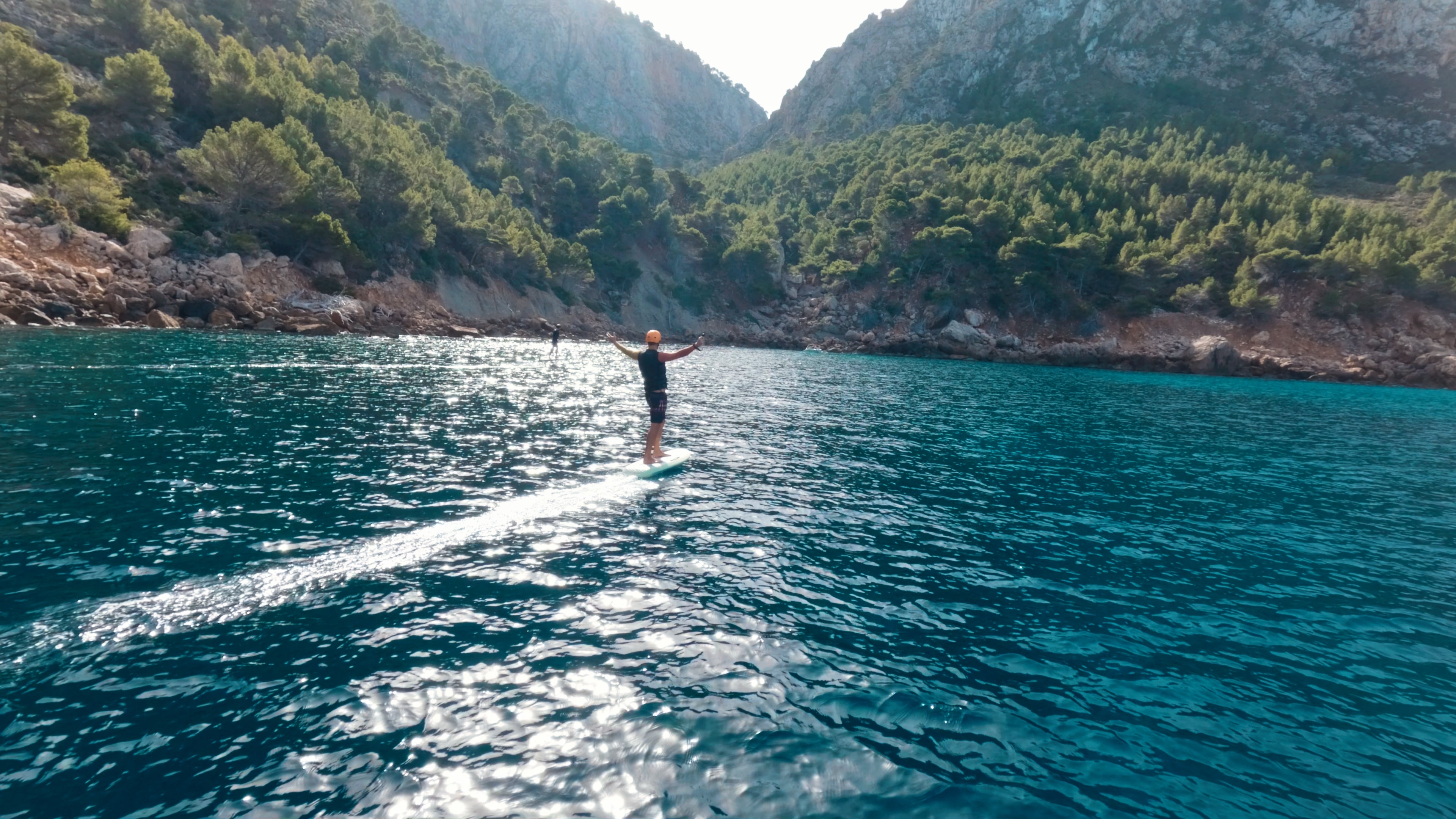Eine Person trudelt auf einem fliteboard auf dem Meer und hält die Hände jubelnd in den Himmel, im Hintergrund sieht man eine andere Person trudeln und mediterrane Berge.