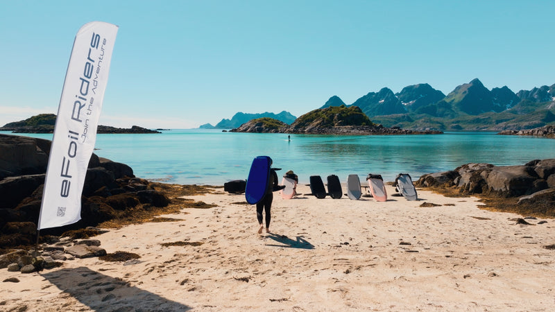 Inmitten der norwegischen Lofoten stehen sechs Fliteboards am Strand und ein Mann trägt das nächste auf seinem Rücken, während im Hintergrund bereits eine Person eFoiling betreibt.
